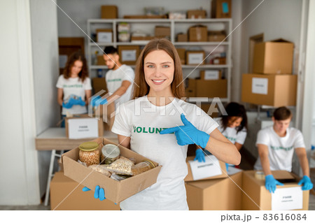Young woman at volunteer center holding and pointing at donations paper box smiling at camera 83616084