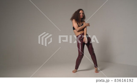 Dancing young African-American girl wearing black top and leather pants make salsa moves with hands in the air, isolated on grey background, emotionally move 83616192