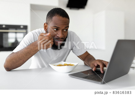 Black man eating cereal at home using laptop 83616383