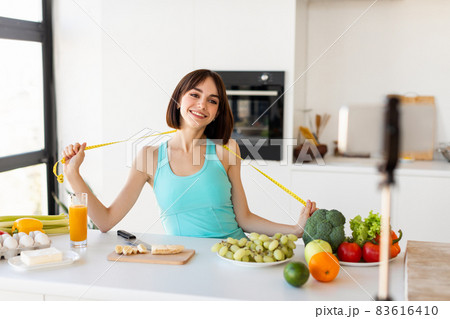 Happy female fitness vlogger filming vlog on smartphone, sharing advice on eating habits, standing with measuring tape 83616410