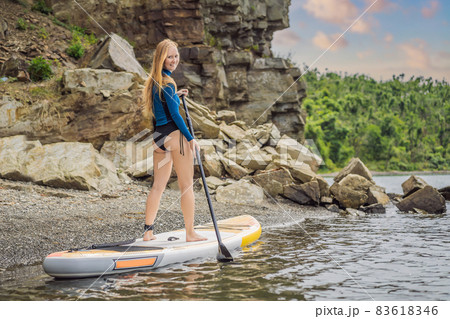 Young women Having Fun Stand Up Paddling in the sea. SUP. Red hair girl Training on Paddle Board near the rocks Young women Having Fun Stand Up Paddling in the sea. SUP. Red hair girl Training on Paddle Board near the rocks 83618346