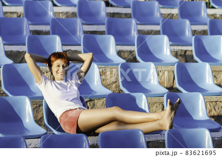 Young woman with short red hair sits relaxed on plastic blue seat in deserted open air audience. Summer vibes. 83621695