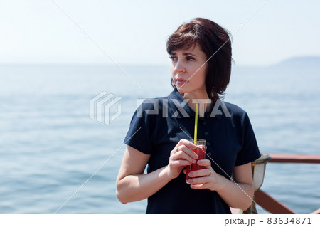 A brunette woman in a cafe on the pier is standing with a glass of juice waiting for the ship A brunette woman in a cafe on the pier is standing with a glass of juice waiting for the ship 83634871