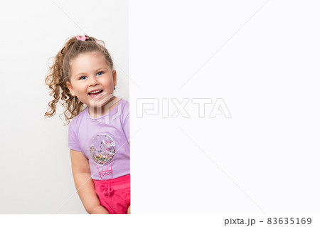 Smiling blue eyed and cute little baby girl peeking behind a white wall. 83635169