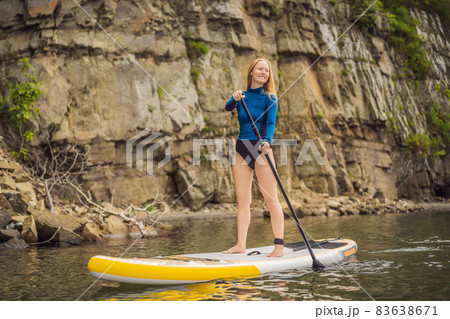 Young women Having Fun Stand Up Paddling in the sea. SUP. Red hair girl Training on Paddle Board near the rocks Young women Having Fun Stand Up Paddling in the sea. SUP. Red hair girl Training on Paddle Board near the rocks 83638671