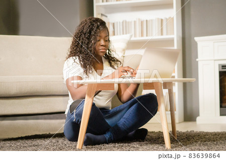 Pretty Afro Girl With Folded Legs Sitting on the Carpet and Typing on her Computer. African Girl Looks Through the List of Films on Her Laptop. Close-up. Home Background Pretty Afro Girl With Folded Legs Sitting on the Carpet and Typing on her Computer. African Girl Looks Through the List of Films on Her Laptop. Close-up. Home Background 83639864