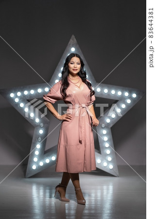 Sexy Asia Milf Model Stands Half a Turn Against the Backdrop of a Giant Star in the Dark Studio. Giant Star with Lights. Full-length. Dark background Sexy Asia Milf Model Stands Half a Turn Against the Backdrop of a Giant Star in the Dark Studio. Giant Star with Lights. Full-length. Dark background 83644381