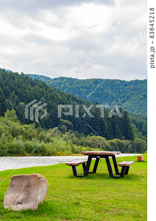 Wooden bench and table on green lawn near river among mountains and dense forest 83645218