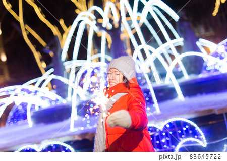 Smiling young woman wearing winter knitted clothes holding sparkler outdoors over snow background. Christmas holidays. Smiling young woman wearing winter knitted clothes holding sparkler outdoors over snow background. Christmas holidays. 83645722