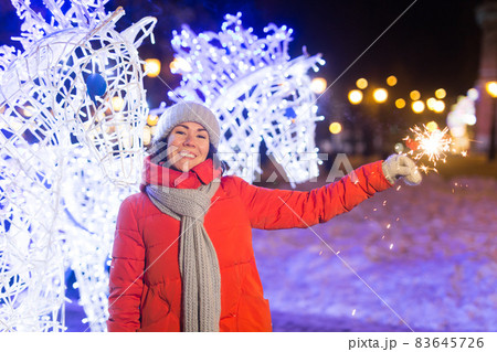 Smiling young woman wearing winter knitted clothes holding sparkler outdoors over snow background. Christmas holidays. Smiling young woman wearing winter knitted clothes holding sparkler outdoors over snow background. Christmas holidays. 83645726