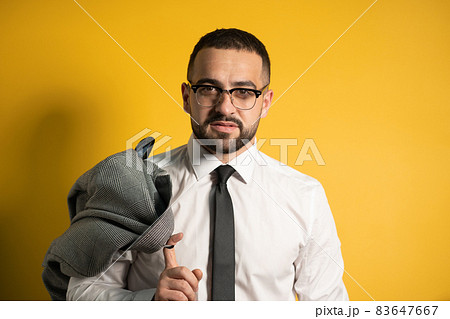 Looking serious business man with trendy beard dressed in greyish suite posing holding his jacket on his shoulder hanging it behind looking isolated on yellow background 83647667