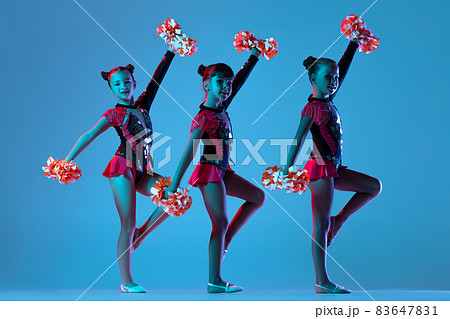 Full-length portrait of three girls, cheerleaders training wearing colorful costumes with pom-pons isolated over blue background in neon lights Full-length portrait of three girls, cheerleaders training wearing colorful costumes with pom-pons isolated over blue background in neon lights 83647831