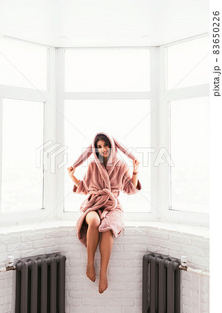 A beautiful woman in a dressing gown is sitting on the windowsill. A woman in a bunny costume. The girl at the window. A pretty girl in a pink robe. 83650226