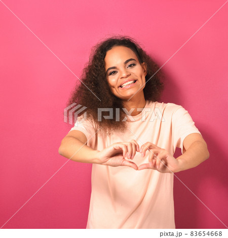 Sending love gesture beautiful young African American woman looking positively at camera wearing peachy t-shirt isolated on pink background. Beauty concept. Square crop 83654668