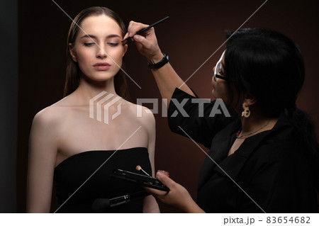 Beautiful model at MUAH artist. Beauty makeup artist working on a fashion model face isolated on black background. Make-up artist, applying eye shadow to the model 83654682