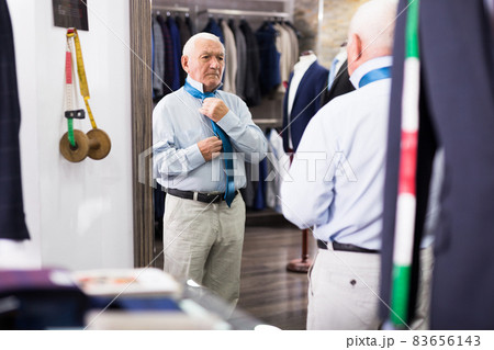 Mature shopper tries on fashionable tie in front of mirror in clothing store 83656143