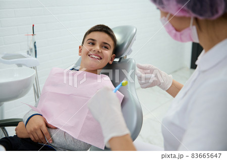 Adorable boy sitting in dentist chair smiles with beautiful toothy smile looking at a pediatric dentist teaching him about oral hygiene during dental appointment in white clean modern medical cabinet Adorable boy sitting in dentist chair smiles with beautiful toothy smile looking at a pediatric dentist teaching him about oral hygiene during dental appointment in white clean modern medical cabinet 83665647