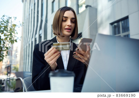Pretty woman, entrepreneur, copywriter holding mobile phone and gold credit card sitting at table in outside public park and working on laptop, shopping online, booking, ticketing. Pretty woman, entrepreneur, copywriter holding mobile phone and gold credit card sitting at table in outside public park and working on laptop, shopping online, booking, ticketing. 83669190