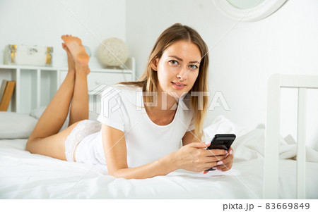 Young woman in lace lingerie lying in bed with mobile phone in hands in morning sunbeams 83669849