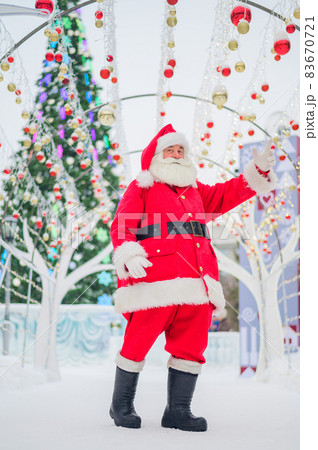 Portrait of an elderly man dressed as santa claus on the background of a christmas tree outdoors. 83670721