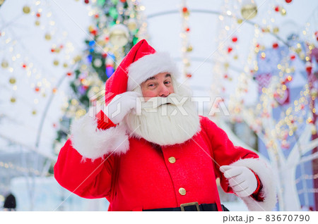 Portrait of an elderly man dressed as santa claus on the background of a christmas tree outdoors. 83670790
