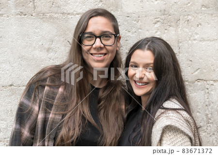 Two young lesbians standing near a stone wall, smiling, happy. Two young lesbians standing near a stone wall, smiling, happy. 83671372