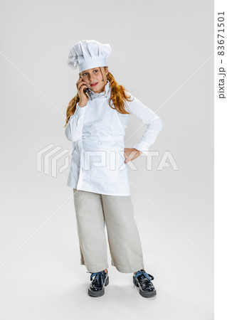 One little cute girl in white cook uniform and huge chef's hat posing isolated on white studio background. 83671501