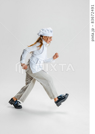 Little Caucasian girl in white cook uniform and huge chef's hat going isolated on white studio background. 83672491