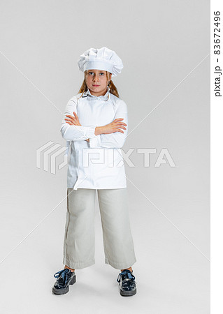 Portrait of little cute girl in white cook uniform and huge chef's hat standing with arms crossed isolated on white studio background. 83672496