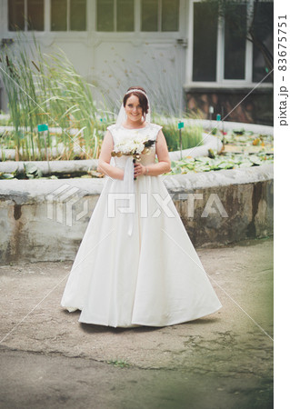 Beautiful brunette bride in elegant white dress holding bouquet posing neat trees 83675751