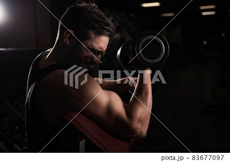 A handsome man with glasses doing an exercise for biceps with a barbell. The guy is engaged in bodybuilding. Trainer in the gym with muscular arms. A handsome man with glasses doing an exercise for biceps with a barbell. The guy is engaged in bodybuilding. Trainer in the gym with muscular arms. 83677097