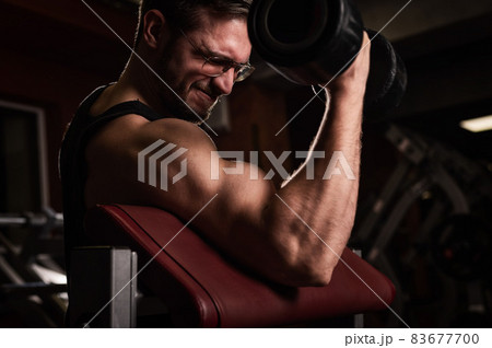 A handsome man with glasses doing an exercise for biceps with a barbell. The guy is engaged in bodybuilding. Trainer in the gym with muscular arms. 83677700