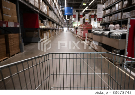 Empty shopping cart in a furniture store warehouse. Supermarket shopping basket aisle with box shelf abstract blur defocused background Empty shopping cart in a furniture store warehouse. Supermarket shopping basket aisle with box shelf abstract blur defocused background 83678742