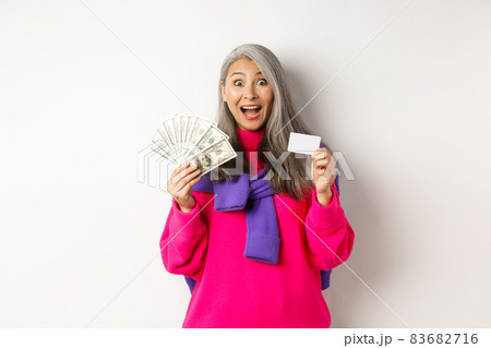 Shopping concept. Amazed asian granny showing plastic credit card and money dollars, want to buy something, standing over white background Shopping concept. Amazed asian granny showing plastic credit card and money dollars, want to buy something, standing over white background 83682716