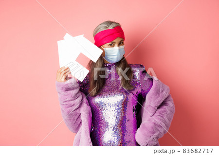Covid-19 and real estate concept. Cool and stylish asian elderly woman in disco dress and medical mask showing paper house, standing over pink background Covid-19 and real estate concept. Cool and stylish asian elderly woman in disco dress and medical mask showing paper house, standing over pink background 83682717