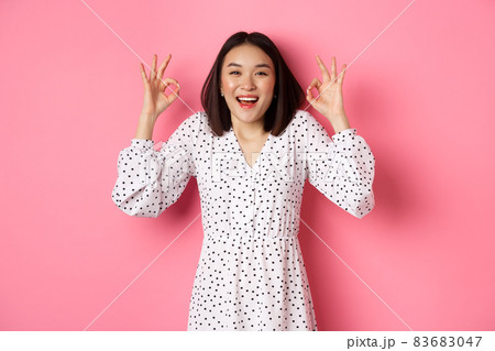 Pretty young asian woman in dress showing okay sign, praising and showing approval, looking satisfied, standing against pink background Pretty young asian woman in dress showing okay sign, praising and showing approval, looking satisfied, standing against pink background 83683047