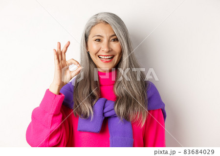 Close up of beautiful asian mature woman showing OK sign and smiling, approve and like product, standing satisfied against white background Close up of beautiful asian mature woman showing OK sign and smiling, approve and like product, standing satisfied against white background 83684029