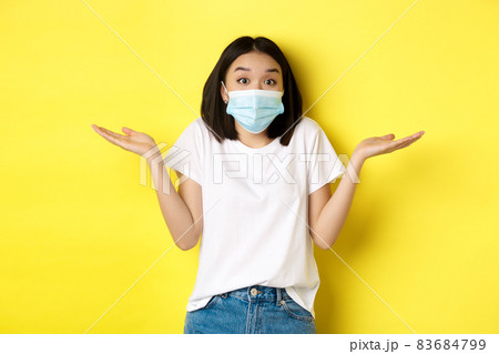 Covid-19, quarantine and social distancing concept. Clueless asian woman in medical mask shrugging shoulders, spread hands sideways clueless, know nothing, yellow background Covid-19, quarantine and social distancing concept. Clueless asian woman in medical mask shrugging shoulders, spread hands sideways clueless, know nothing, yellow background 83684799