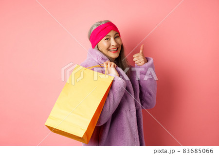 Stylish asian senior woman going shopping, carry paper bag on shoulder and showing thumb-up, recommending store discounts, pink background Stylish asian senior woman going shopping, carry paper bag on shoulder and showing thumb-up, recommending store discounts, pink background 83685066