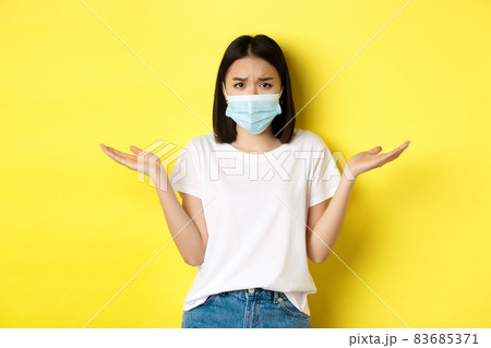 Covid-19, quarantine and social distancing concept. Confused asian woman in medical mask shrugging shoulders, spread hands sideways clueless, know nothing, yellow background Covid-19, quarantine and social distancing concept. Confused asian woman in medical mask shrugging shoulders, spread hands sideways clueless, know nothing, yellow background 83685371