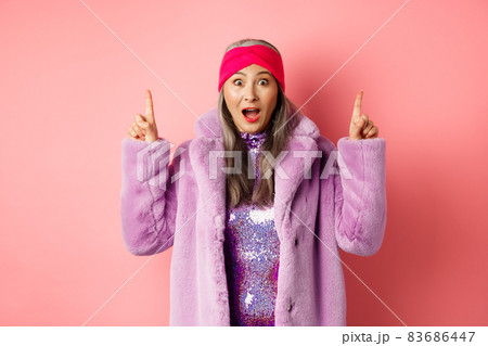 Happy elderly asian lady pointing fingers up, looking amazed at camera, showing cool promo offer, standing against pink background in purple fake fur coat 83686447