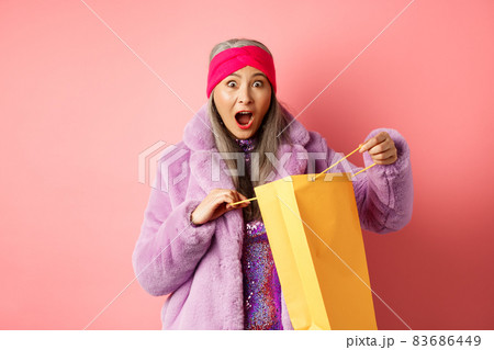 Shopping and fashion concept. Surprised asian grandmother open shop bag and looking amazed at camera, receive mother day gift, pink background 83686449
