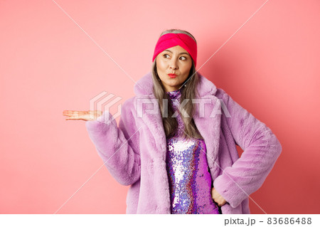 Fashion and shopping concept. Stylish asian senior woman in purple winter outfit demonstrating product on hand. Cool old lady in trendy clothes holding something on palm, pink background Fashion and shopping concept. Stylish asian senior woman in purple winter outfit demonstrating product on hand. Cool old lady in trendy clothes holding something on palm, pink background 83686488