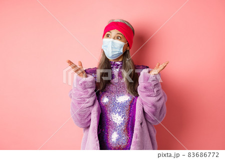 Covid-19, social distancing and fashion concept. Surprised and shocked asian stylish granny looking left, spread hands sideways confused, standing over pink background 83686772
