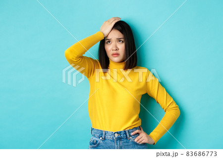 Tired and sad asian woman facepalm, sighing and looking up distressed, feeling troubled while standing against blue background 83686773