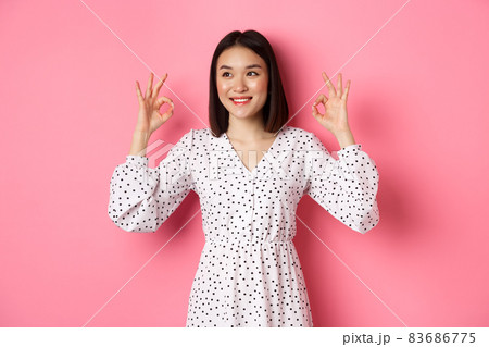 Pretty young asian woman in dress showing okay sign, praising and showing approval, looking satisfied, standing against pink background Pretty young asian woman in dress showing okay sign, praising and showing approval, looking satisfied, standing against pink background 83686775