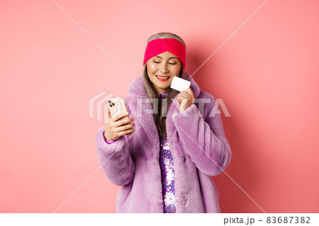 Online shopping and fashion concept. Stylish asian grandmother making purchase in internet with smartphone and plastic credit card, pink background Online shopping and fashion concept. Stylish asian grandmother making purchase in internet with smartphone and plastic credit card, pink background 83687382