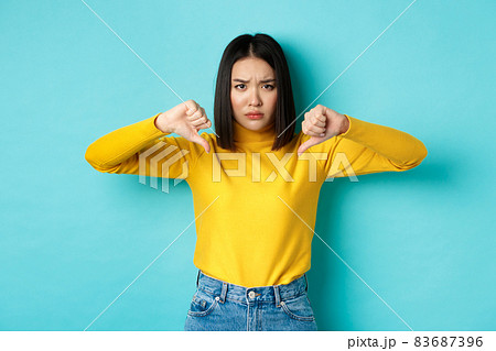 Disappointed asian woman frowning upset, showing thumbs down in dislike and disapproval, standing over blue background, staring at camera 83687396