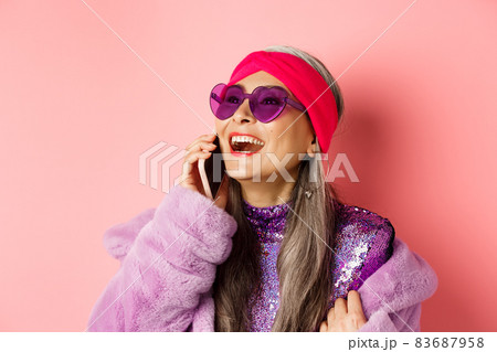 Fashion. Beautiful asian senior woman in trendy sunglasses and headband laughing, talking on mobile phone and looking aside, pink background Fashion. Beautiful asian senior woman in trendy sunglasses and headband laughing, talking on mobile phone and looking aside, pink background 83687958