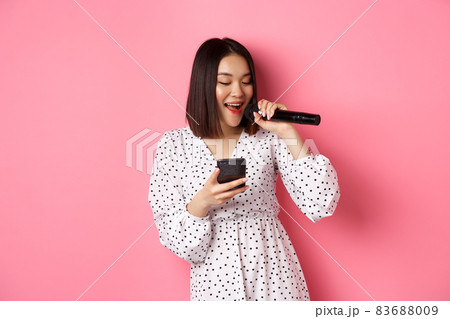 Cute asian woman reading lyrics in smartphone, singing with microphone, standing in trendy dress over pink background Cute asian woman reading lyrics in smartphone, singing with microphone, standing in trendy dress over pink background 83688009